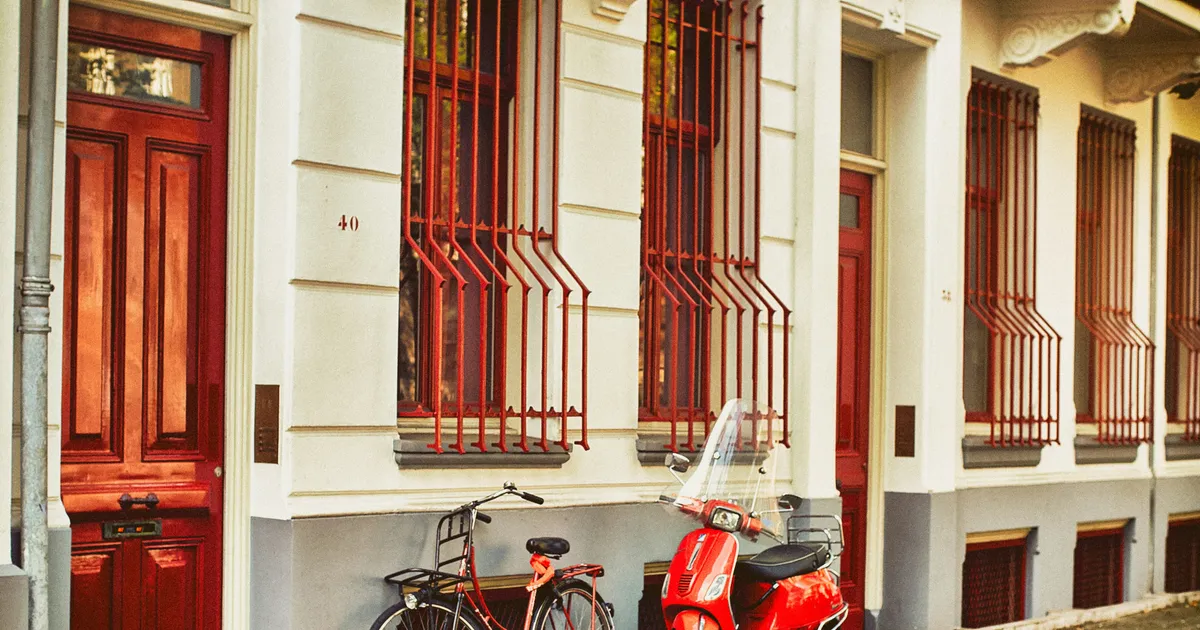 Een rode scooter en een fiets staan naast elkaar tegen de gevel van een herenhuis