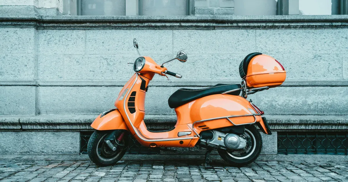 Een oranje scooter staat geparkeerd op een straat met kinderkopjes voor een stenen gevel